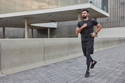Determined bearded adult man in activewear jogs in city street covers long distance enjoys morning running exercises outdoors keeps fit and healthy muscular strength flexibility physical power