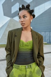 Fashionable woman with bun hairstyle dressed in stylish green clothes keeps hands in pocket looks away pensively poses at urban place during daytime. People street style and lifestyle concept