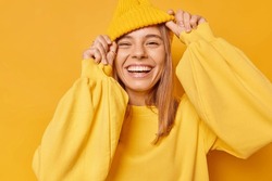 Happy playful young woman smiles broadly shows white teeth keeps hands on hat dressed in casual jumper expresses sincere emotions being in good mood isolated over yellow background. Joy concept
