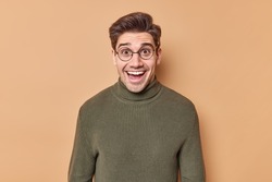 Excited cheerful young man smiles happily and looks with surprised expression reacts on awesome news wears spectacles and sweater isolated over beige background glad to see company of friends