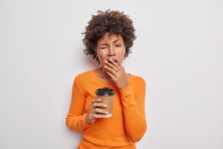 Waist up shot of tired sleepy curly brunette woman covers mouth and yawns drinks caffeine beverage to refresh wears casual orange jumper isolated over white background. People and tiredness concept