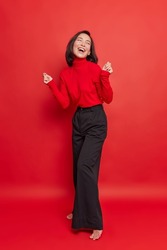 Full length shot of beautiful brunette woman dances carefree keeps hands raised wears turtleneck and black trousers isolated over vivid red background. Energetic carefree female model poses indoor