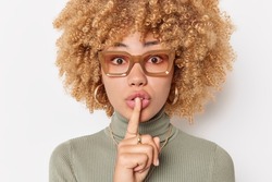 Mysterious curly haired young woman presses index finger over lips makes silence gesture asks to be quiet wears spectacles and turtleneck isolated over white background. Hush be quiet please