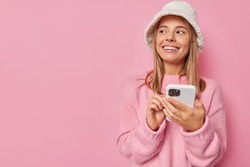 Photo of smiling European woman looks with happy expression uses mobile phone for chatting online wears winter panama and cashmere jumper isolated over pink background copy space for promotion
