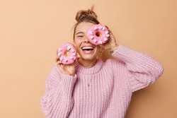 Happy joyful woman covers eye with delicious glazed doughnut laughs happily dressed in casual knitted sweater poses indoor against beige background has sweet tooth breaks diet. Junk food concept
