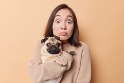 Indoor shot of impressed young Asian woman poses with pug dog shocked to finds out something embarrassing sees something breathtaking poses against brown background. Domestic animals concept