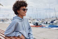 Sideways shot of positive curly haired woman poses on bench wears sunglasses and casual blue hoodie smiles broadly enjoys beautiful view of harbor breathes fresh marine air rests after walking