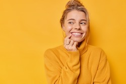 Beautiful glad woman smiles tenderly looks away with happy expression thinks about pleasant things wears casual soft jumper isolated over yellow background blank copy space for your promotion