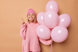 Making wish concept. Positive birthday girl crosses fingers believes in miracle happen holds bunch of inflated balloons wears hat and casual jumper isolated over beige background celebrates event
