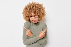 Hesitant clueless woman with curly hair points at different sides picks two variants hesitates about something chooses between wears eyeglasses and turtleneck isolated over white background.