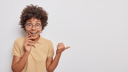 Happy surprised young woman with curly hair points thumb away shows amazing news demonstrates promotion dressed in casual wear isolated over white background. People and advertisement concept