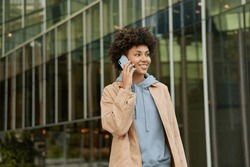 Horizontal shot of happy young woman makes telephone call in roaming wears casual sweatshirt and beige jacket had glad expression walks against blurred city building enjoys smartphone talking