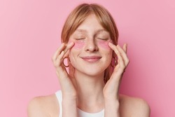 Pleased redhaired female model undergoes beauty procedures appies hydrogel patches under eyes to remove puffiness and fine lines wears casual t shirt isolated over pink background. Skin care