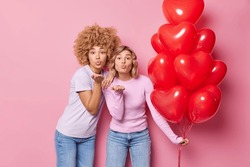 Romantic affectionate two women send air kiss hold bunch of heart balloons dressed ccasually celebrates Valentines Day isolated over pink background. People friendshipp and celebration concept