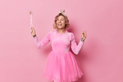 Positive overjoyed woman entertains people on holiday pretends being fairy wears dress holds magic wand laughs gladfully isolated over pink background. People celebration and partying concept