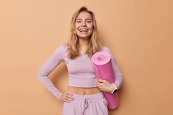 Cheerful woman leads sporty lifestyle dressed in activewear hold rolled mat being in good mood prepares for training in gym smiles broadly isolated over brown background. Regular workout concept
