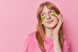 Horizontal shot of dreamy teenage girl with natural red hair puts on green hydrogel patches under eyes for skin treatment dressed in casual t shirt isolated over pink background blank space.