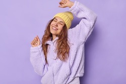 Waist up shot of carefree young European woman with long hair daces and catches every bit of music smiles broadly keeps eyes closed dressed in winter outerwear isolated over purple background