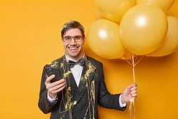 Positive man in festive clothes celebrates anniversary holds bunch of helium balloons and smartphone wears festive clothes being in good mood gets congratulations isolated over yellow background
