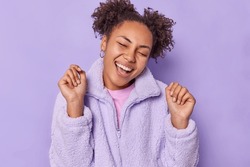 Optimistic cheerful teenage girl keeps eyes closed tilts head shakes hands feels full of happiness rejoices pleasant news wears winter fur jacket isolated over purple background. Emotions concept