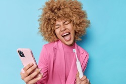 Positive carefree woman with curly bushy hair holds electric brush as if microphone makes selfie photo via smartphone dressed in formal pink jacket isolated over bue background. Hygiene procedures