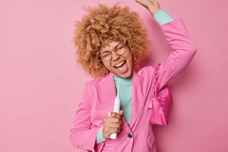 Glad overjoyed curly haired young woman holds electric toothbrush as if microphone sings song dressed in fashionable formal outfit has happy mood dances carefree against pink background has fun
