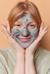 Vertical shot of pretty satisfied millennial foxy girl keeps hands near cheeks applies nourishing clay mask for skin treatment looks directly at camera poses indoor. Beauty and wellness concept