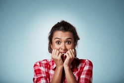 Close-up portrait of a young woman scared ,afraid and anxious biting her finger nails, looking at camera with wide opened eyes isolated on a blue background. Human emotions