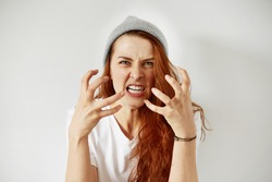 Close up isolated portrait of young annoyed angry woman holding hands in furious gesture. Young female with red hair in white T-shirt and cap. Negative human emotions, face expressions. Film effect 
