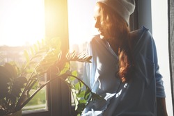 Portrait of happy Caucasian female student spending summer holidays in a big city. Hipster teenager with red hair in gray cap and shirt looking and smiling through the window. Flare sun, film effect 