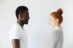 Sideways portrait of a happy couple staring at each other in white morning room. Afro Amercian man in white shirt and redhead girl in stripped longsleeve smiling, showing positive emotions and joy.
