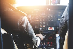 Two unrecognizable jet airliner pilots wearing leather jackets piloting aircraft at sunset, sitting inside cabin at steering control with modern dashboard, sun shining through cockpit windscreen