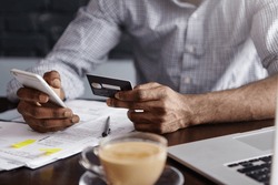 Close up shot of African man's hands holding mobile and credit card. Dark-skinned businessman checking account balance, using online banking application on his cell phone, drinking coffee at cafe