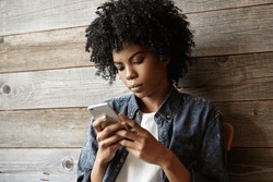 Portrait of beautiful young dark-skinned female with Afro haircut wearing stylish jeans shirt using high-speed Internet connection on generic smart phone, typing messages to her friends online
