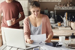 Happy beautiful woman reading notification from bank on prolongation of mortgage term while working through papers at kitchen table with laptop and calculator, her husband standing behind her