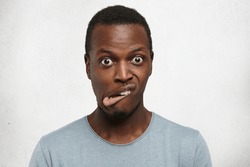 Indoor shot of emotional young dark-skinned man in grey t-shirt grimacing, bugging eyes out, making goofy faces, twisting lips, looking at camera with crazy and stupid expression on his face