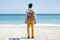 Rear shot of stylish young Afro American backpacker standing on boardwalk on pebble beach, facing vast calm ocean with clear azure water during peaceful morning, admiring amazing marine view