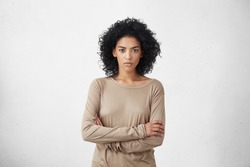 Indoor shot of angry grumpy young mixed race female dressed casually keeping arms folded, looking at camera with strict and skeptical expression, doesn't believe excuses of her unfaithful boyfriend