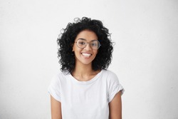 Pretty female with dark skin and broad happy smile wearing big round eyeglasses enjoying good positive news concerning her promotion at work, posing isolated against white blank wall background