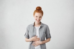 Happy cheerful young Caucasian red haired female with bun smiling cheerfully, keeping hands on her stomach, feeling full after dinner or can't stop laughing at some really good joke. Body language