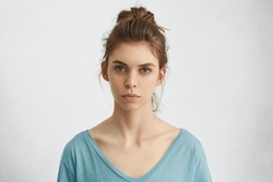 Portrait of young European female with healthy clean skin and blue eyes wearing casual top looking at camera with serious expression. Caucasian woman model with hair gathered in bunch posing indoors