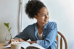 Mixed race woman with crisp hair, dark skin, wearing jean jacket looking aside while sitting at chair, reading her favourite book and drinking coffee, People, hobby, recreation, lifestyle concept