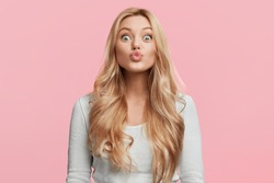 Portrait of adorable blonde woman pouts lips and foolishes alone, makes grimace, rounds lips for making kiss, isolated over pink background. Facial expressions, human emotions and beauty concept