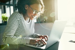 Smiling young African woman wearing glasses working online with a laptop while sitting alone at a table