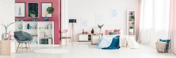 Grey armchair and pink wall in spacious bedroom interior with blue blanket on bed next to a wooden lamp