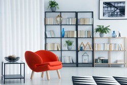 Orange armchair next to table in bright living room interior with bookshelf and poster on white wall