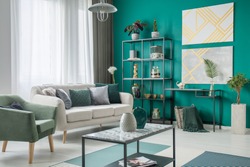 Bright sofa with many pillows standing next to a green armchair in living room interior with metal furniture and geometric paintings