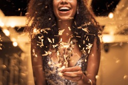 Cheerful young woman holding single sparkler in hand outdoor. Detail of african girl celebrating new year’s eve with bengal light. Closeup of beautiful woman holding a sparkling stick at party night.
