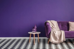 Dark purple sofa with a blanket beside a small table with bottles standing on black and white checkerboard floor in a minimalistic living room interior. Copy space. Real photo
