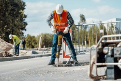 Worker in reflective vest with drill repairing street during roadworks